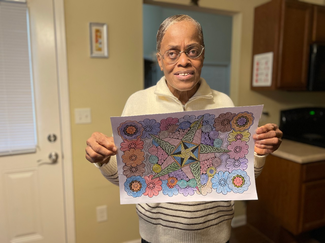 an older black woman with glasses and her gray hair pulled back from her face stands in a kitchen holding up a very detailed colored in sketch of a mandala with patterns and flowers and smiles for the camera