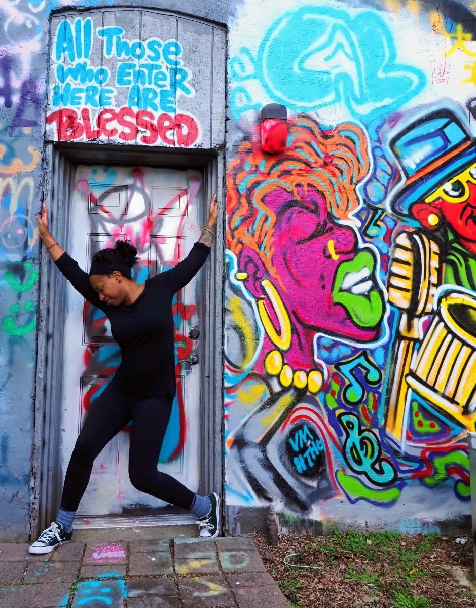 a photo of a young black woman in a dance pose in a doorway with her head bowed to her right and slightly smiling, her arms outstretched and hands braced against the doorframe, and legs slightly bent at an angle; she is shown outdoors against a wall full of vibrant colorful graffiti, including a phrase above the door that says "all that enter here are blessed."  the pavement at her feet in covered in colorful chalk drawings