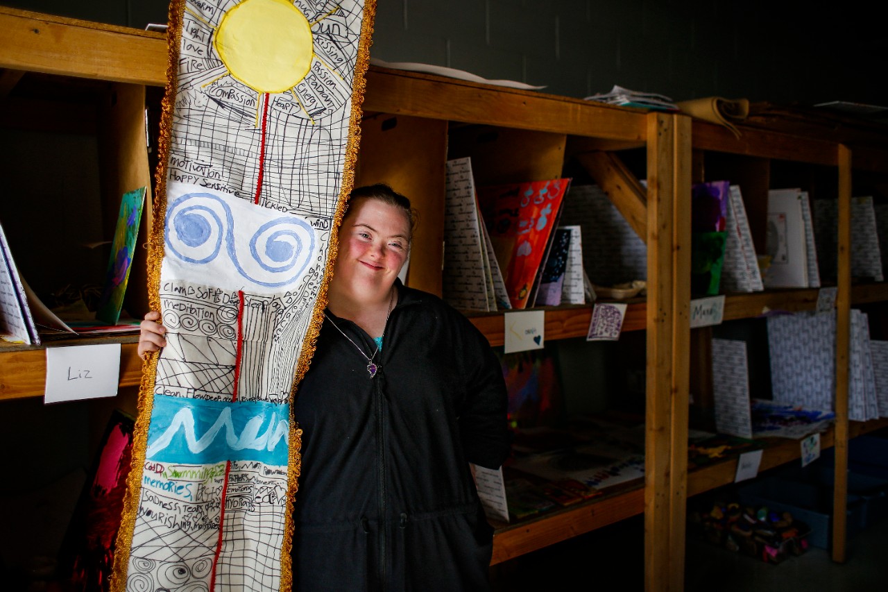 a young white woman with down syndrome stands in front of some wooden shelves holding on to a large 3-D textile work of art that appears to include fabric and drawings showing the sun and other natural shapes