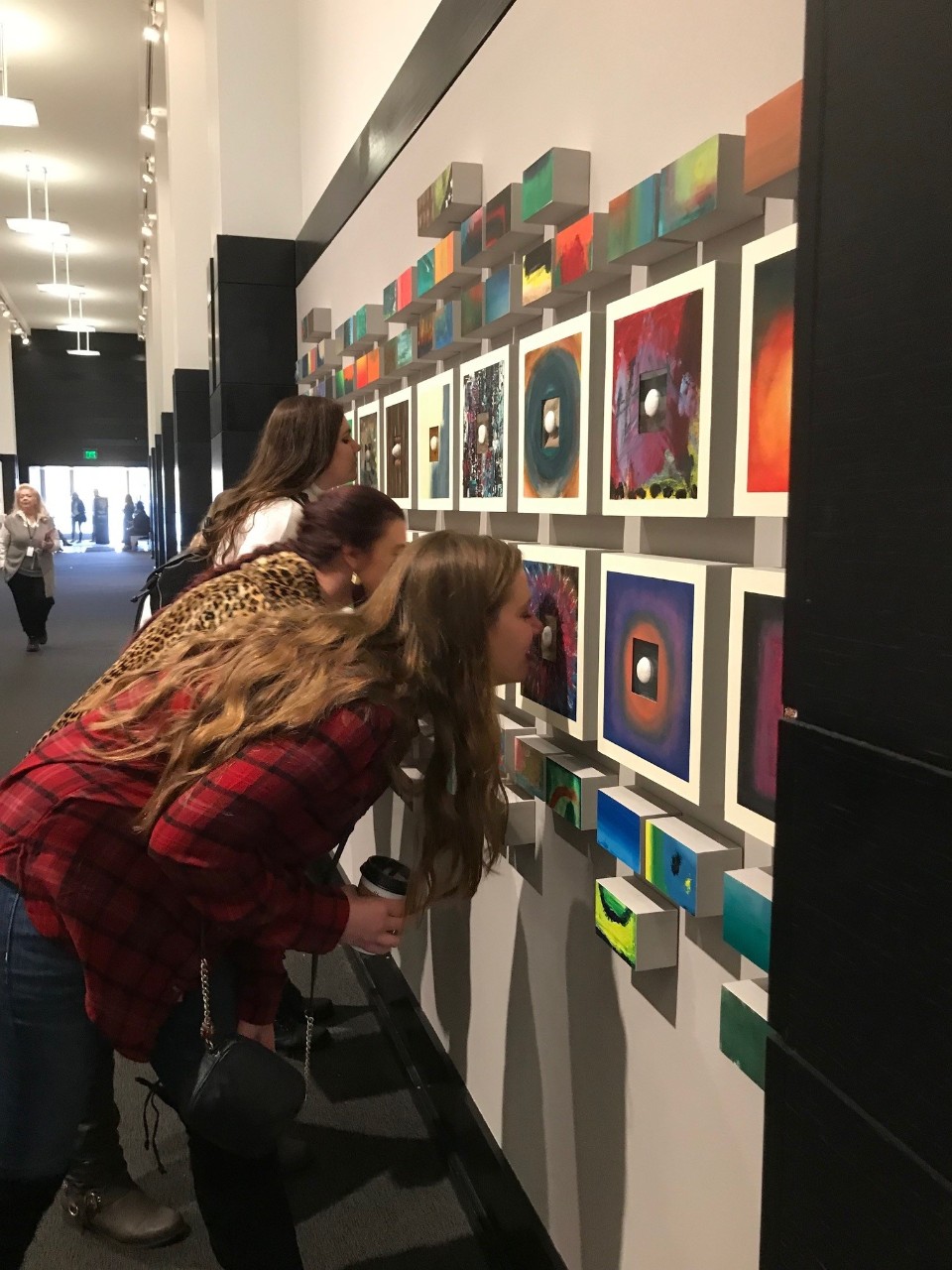 several young women lean close to a display of several colorful textile squares mounted on a wall