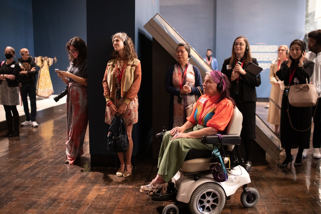 several patrons, including a woman in a power wheelchair (Alicia Searcy), gather in the museum with headphones on to listen to the audio tour of the artwork