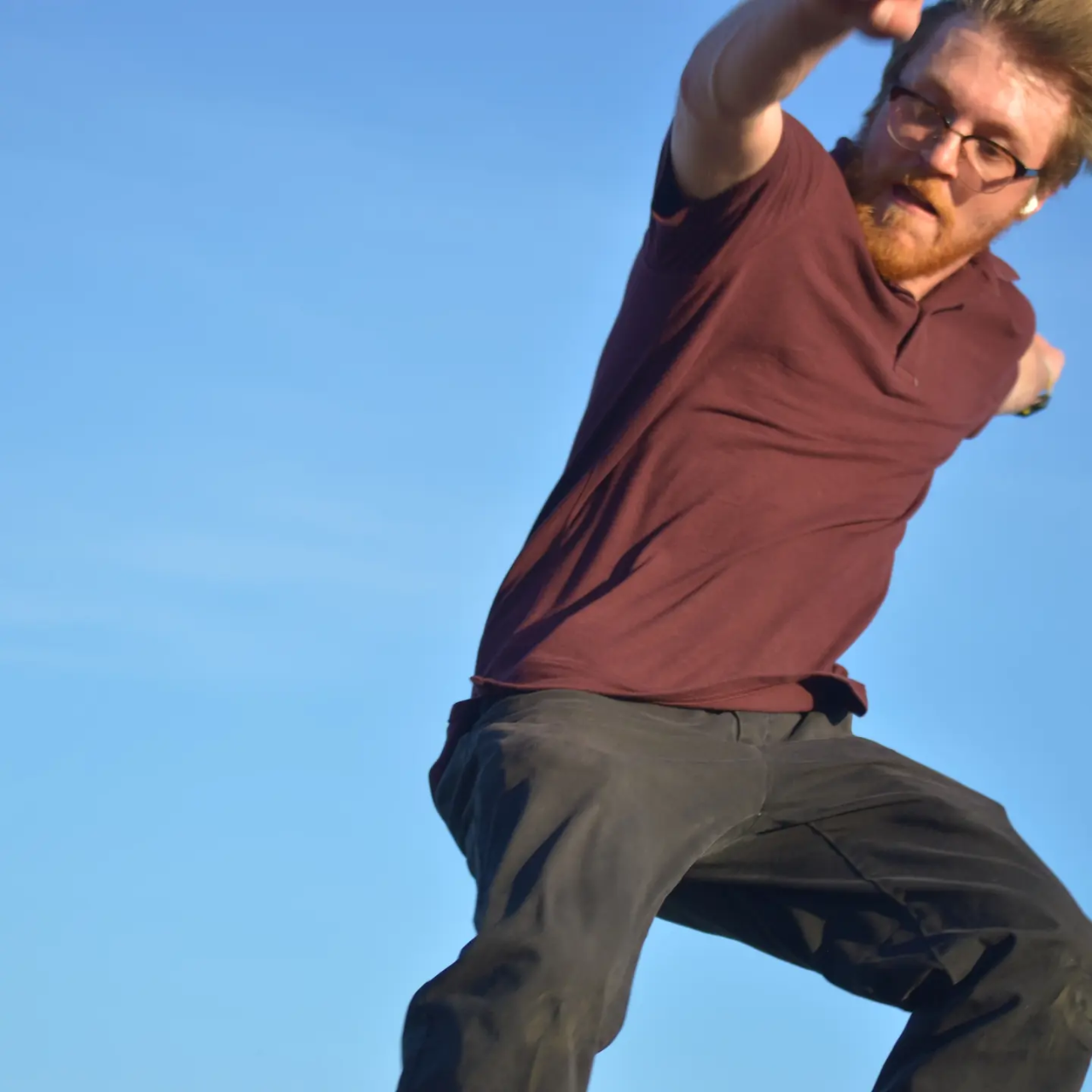 a young man with reddish hair, a beard, glasses and a maroon shirt is shown in the photo jumping up in the air, with a look of concentration on his face as he looks back at the ground. It almost looks like he is in the middle of a skateboard trick but the photo is tight, showing just his body mid-jump