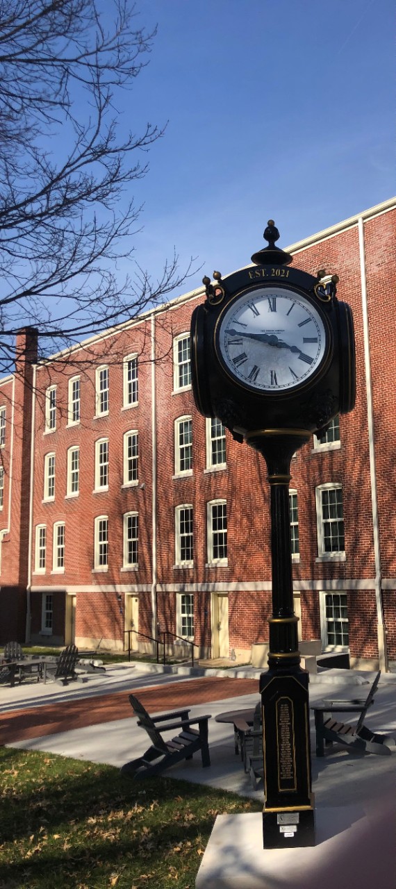 a photo of an ornate black clock on top of a pole, similar to a lamppost, on the ETSU campus with picnic style seating and a brick building in the background