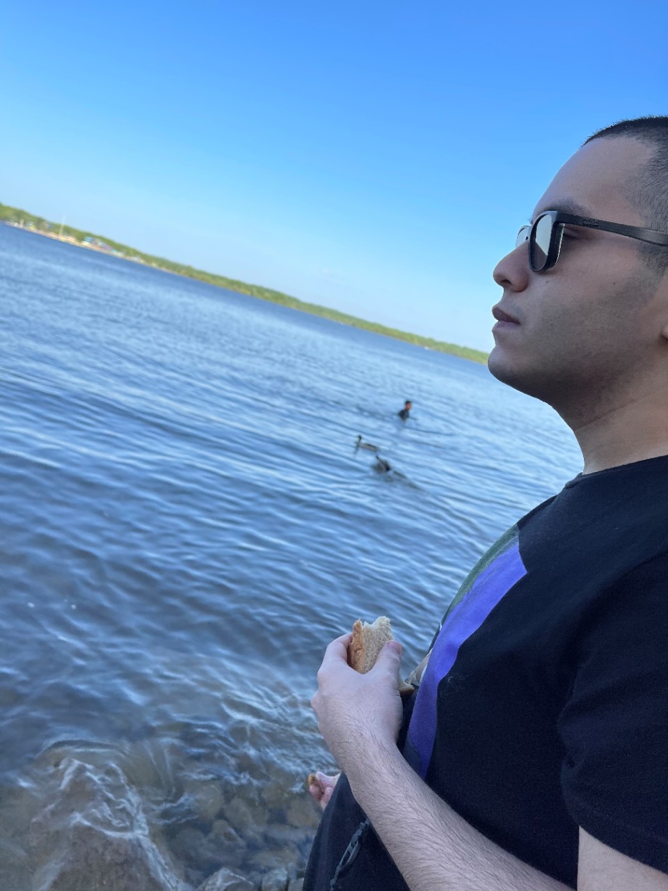 a young Latino man standing by a body of water, wearing a black tshirt and sunglasses