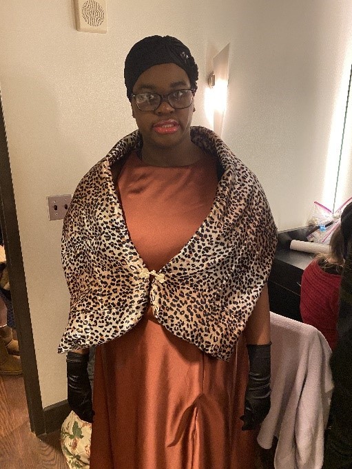 a young black woman with a black head covering, a brown silky long dress, black fancy gloves, a cheetah-print wrap and stage makeup