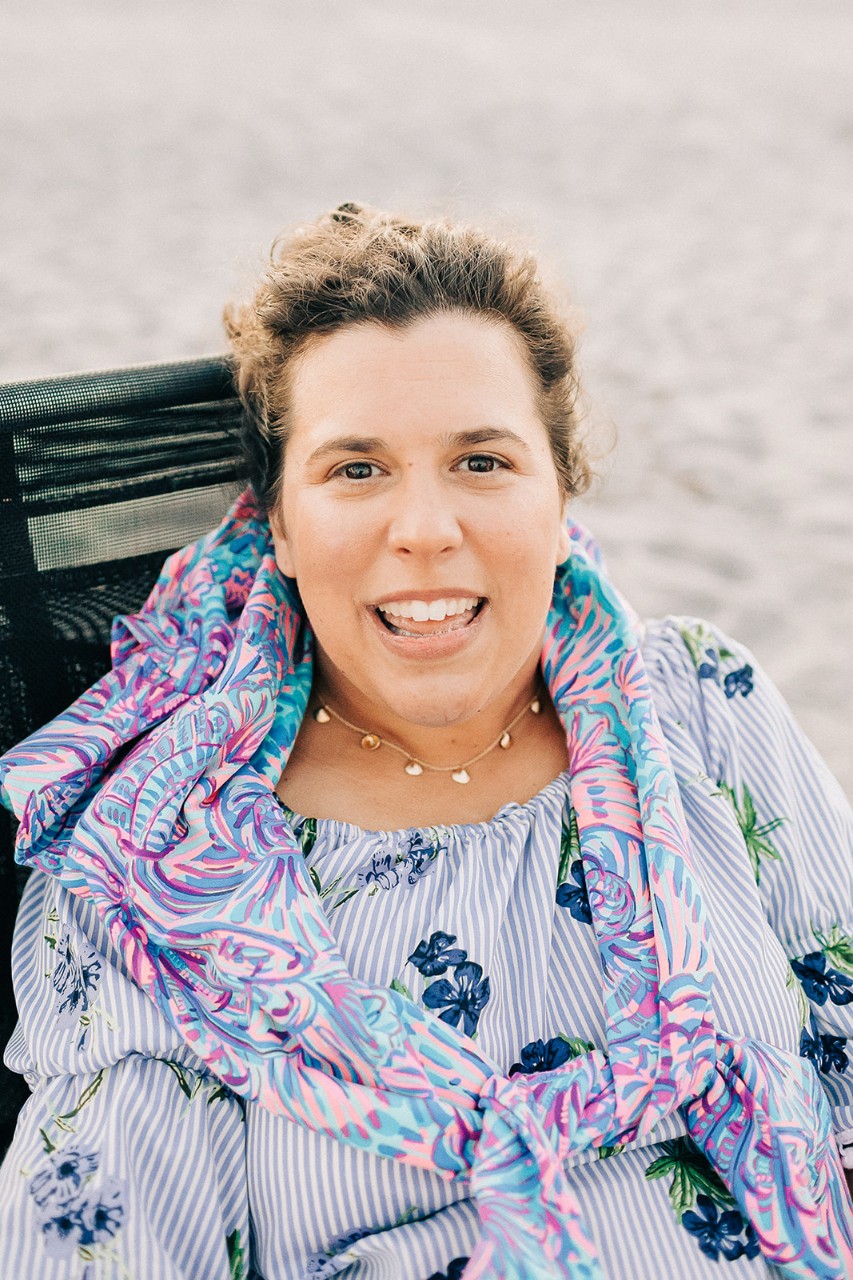 a portrait of a young white woman with short dark blonde hair sitting in her wheelchair on a sandy beach, wearing a colorful scarf and blue flowered blouse and a pretty silver necklace