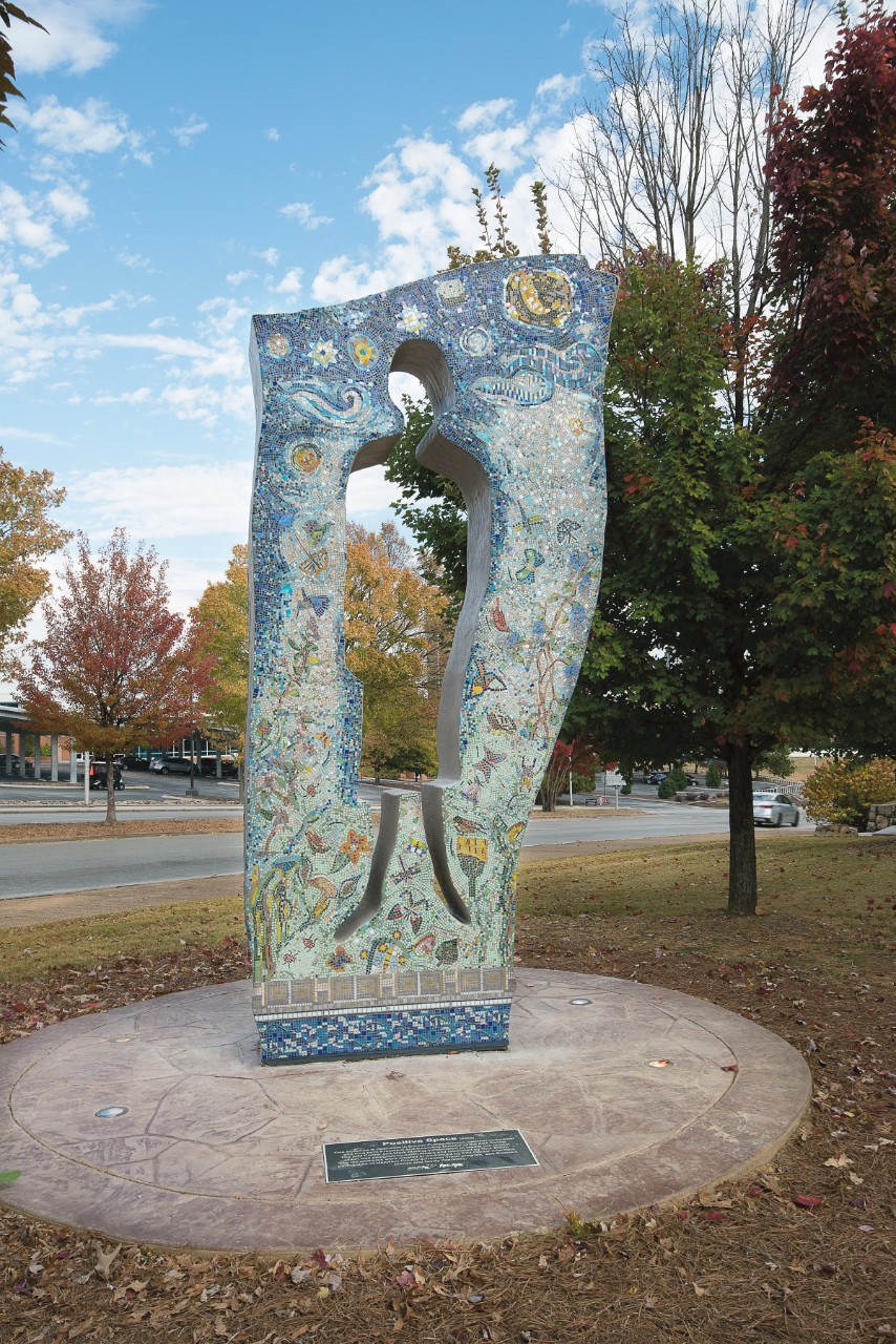 a tall outdoor mosaic sculpture that surrounds an empty space of a human body – head, body, legs – and the surrounding mosaic tiles are in varying shades of blue and many other colors in the shapes of suns, moons, stars, butterflies, dragonflies, and similar figures