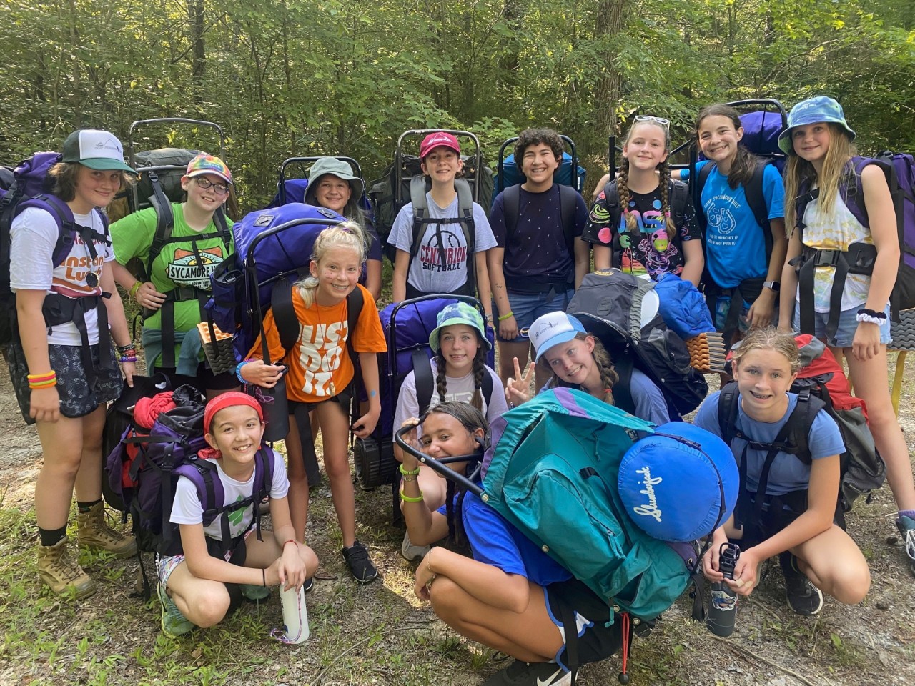 a photo of a diverse group of about 15 young girls in shorts, T-shirts and camping gear like boots and backpacks posing together in the woods and smiling