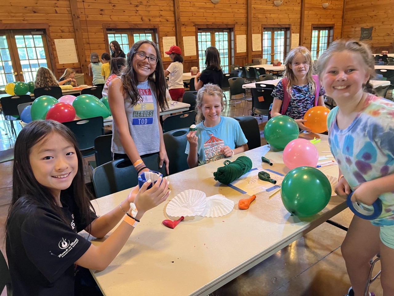 a group of 5 young girls sit at a table in a large cabin with markers and balloons and smile for the camera