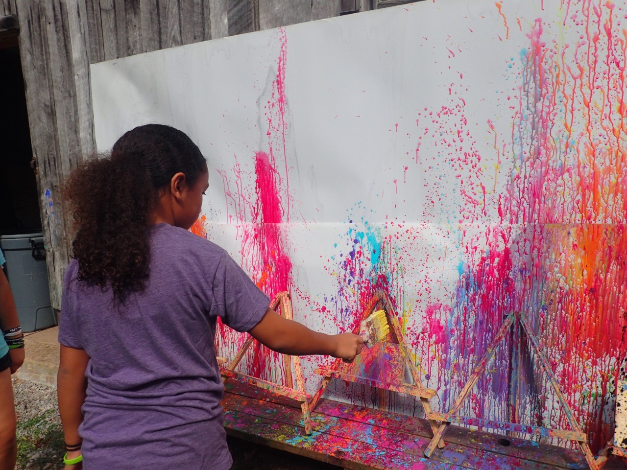 a photo of a young black girl with a purple shirt and a long ponytail holding a paintbrush in front of a huge piece of paper on a wall splattered with bright paint in pinks, oranges, reds and purples