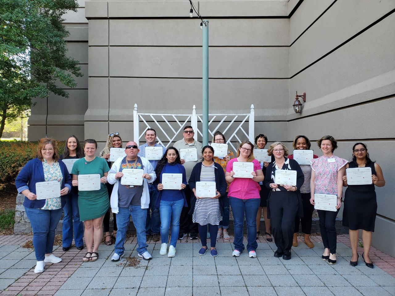 a diverse group of about 20 adults with and without disabilities pose for a group photo in the hotel courtyard holding up their Partners paper certificates.