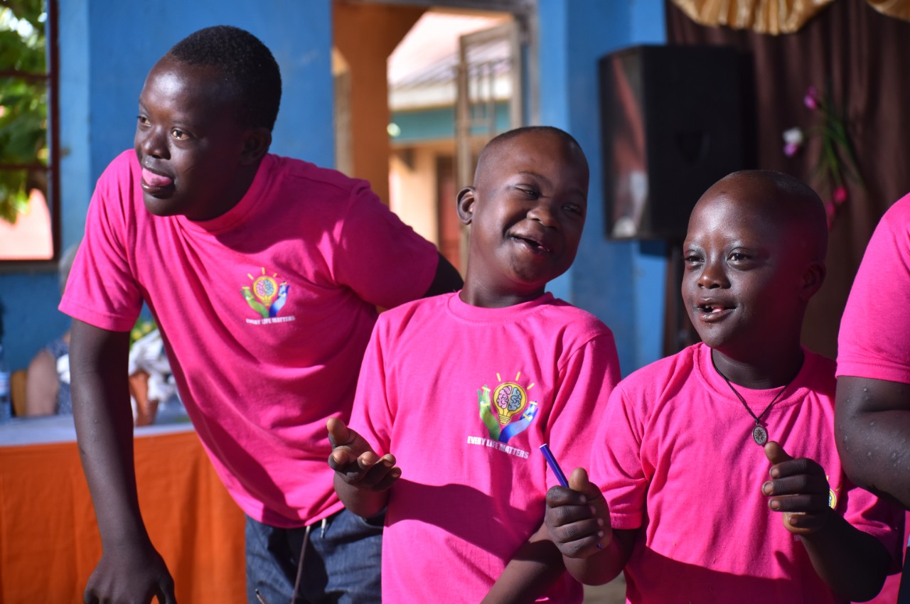Three young Ugandan boys of varying ages who all have Down syndrome are pictured with joyful smiles or in the midst of laughing and matching bright pink shirts