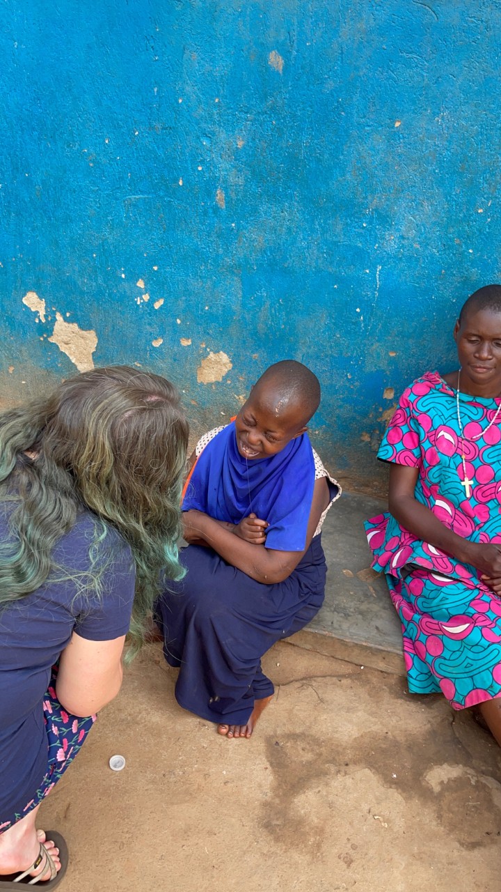 Photo shows Jolene crouching down to speak with two young Ugandan girls who are seated against a bright blue painted wall outside in the dirt, and they are smiling