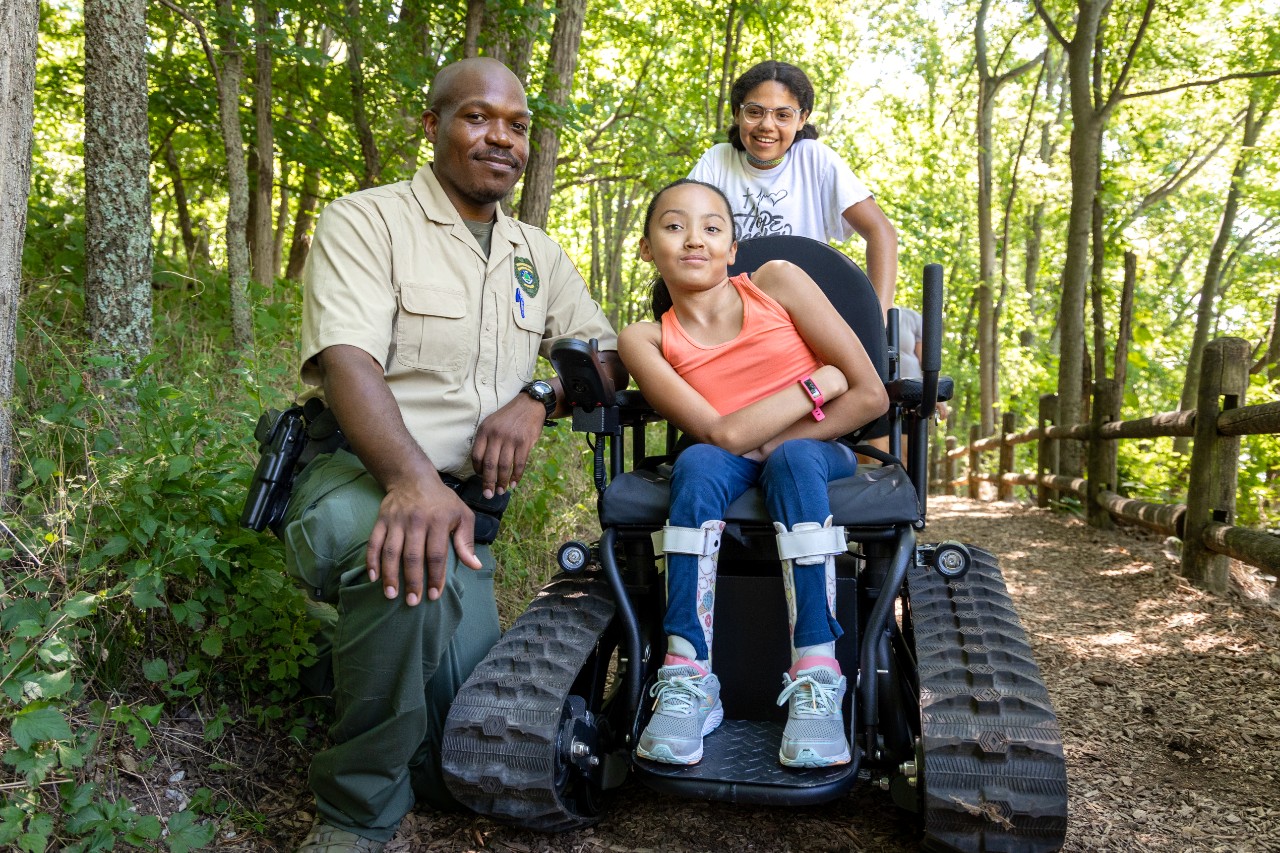 a park ranger is shown kneeling outside in the woods on a nature trail in his uniform next to a young girl seated in an all terrain wheelchair with giant wheel treads; she is in athletic clothes and smiling, and has braces on her legs. another girl about her age, possibly her sister, stands behind the wheelchair smiling too.