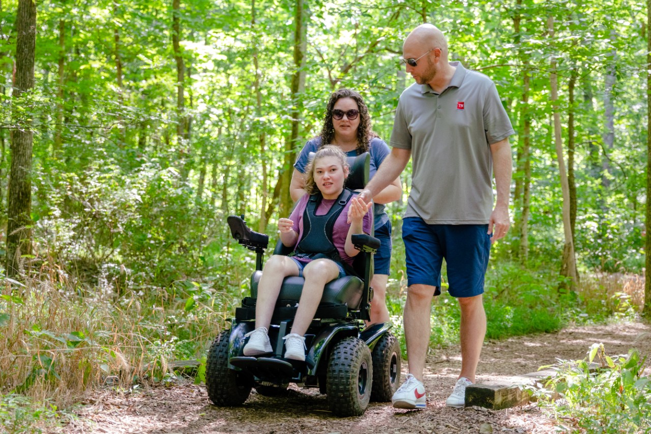 DIDD Commissioner Brad Turner, a white man with athletic clothes on, stands next to his wife Rebecca on a nature trail at Radnor Lake park with their daughter Kinsley, a teenage wheelchair user, in an all terrain wheelchair