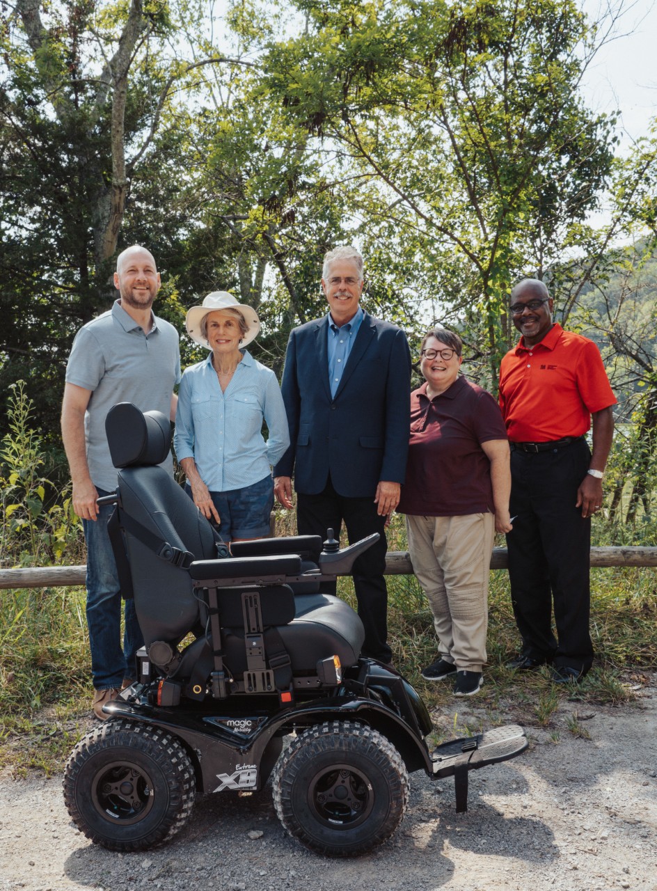 a group of people stand in front of the all-terrain wheelchair in an outdoor photo.
