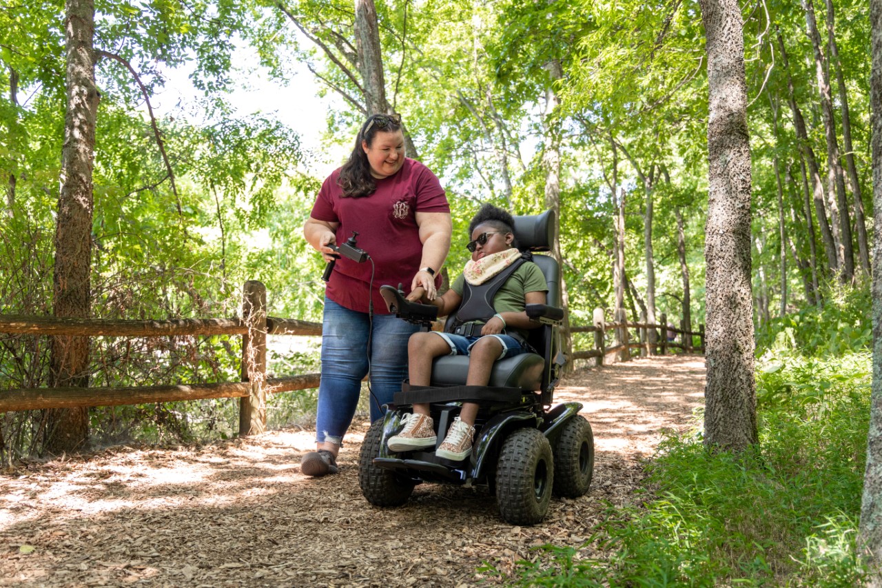 Mom Alison is smiling and walking next to her daughter Charlotte, helping Charlotte use the steering controls of the all-terrain wheelchair on a nature trail.