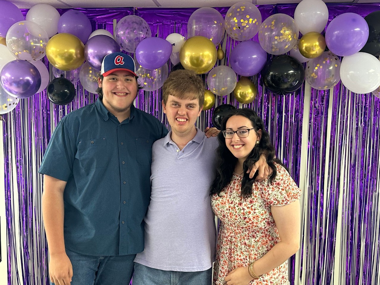 three white, young adults pose with their arms around each other in front of a purple drape background with purple, black, and gold balloons 