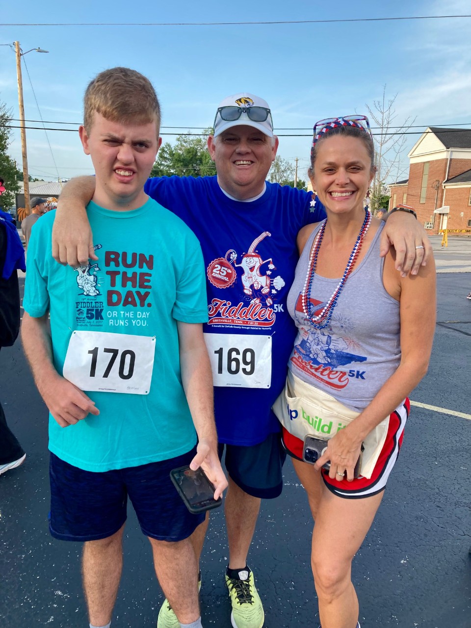 A white young man and two older adults pose together, smiling at the camera. They are wearing race bibs and Fourth of July garb. 