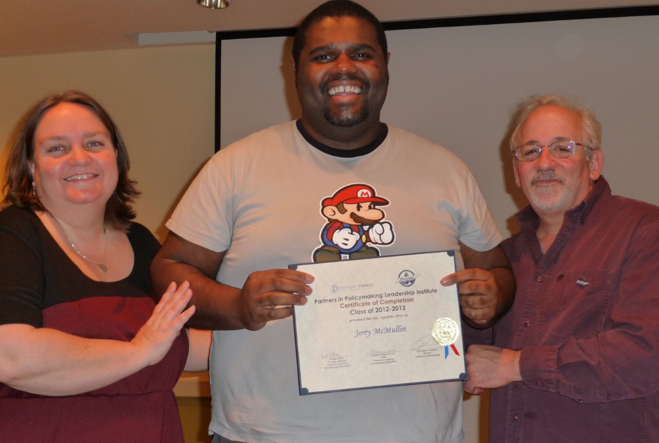 A white woman with shoulder-length brown hair, a Black man in a Mario shirt holding a certificate, and a white man with gray hair and beard and glasses smile at the camera.  