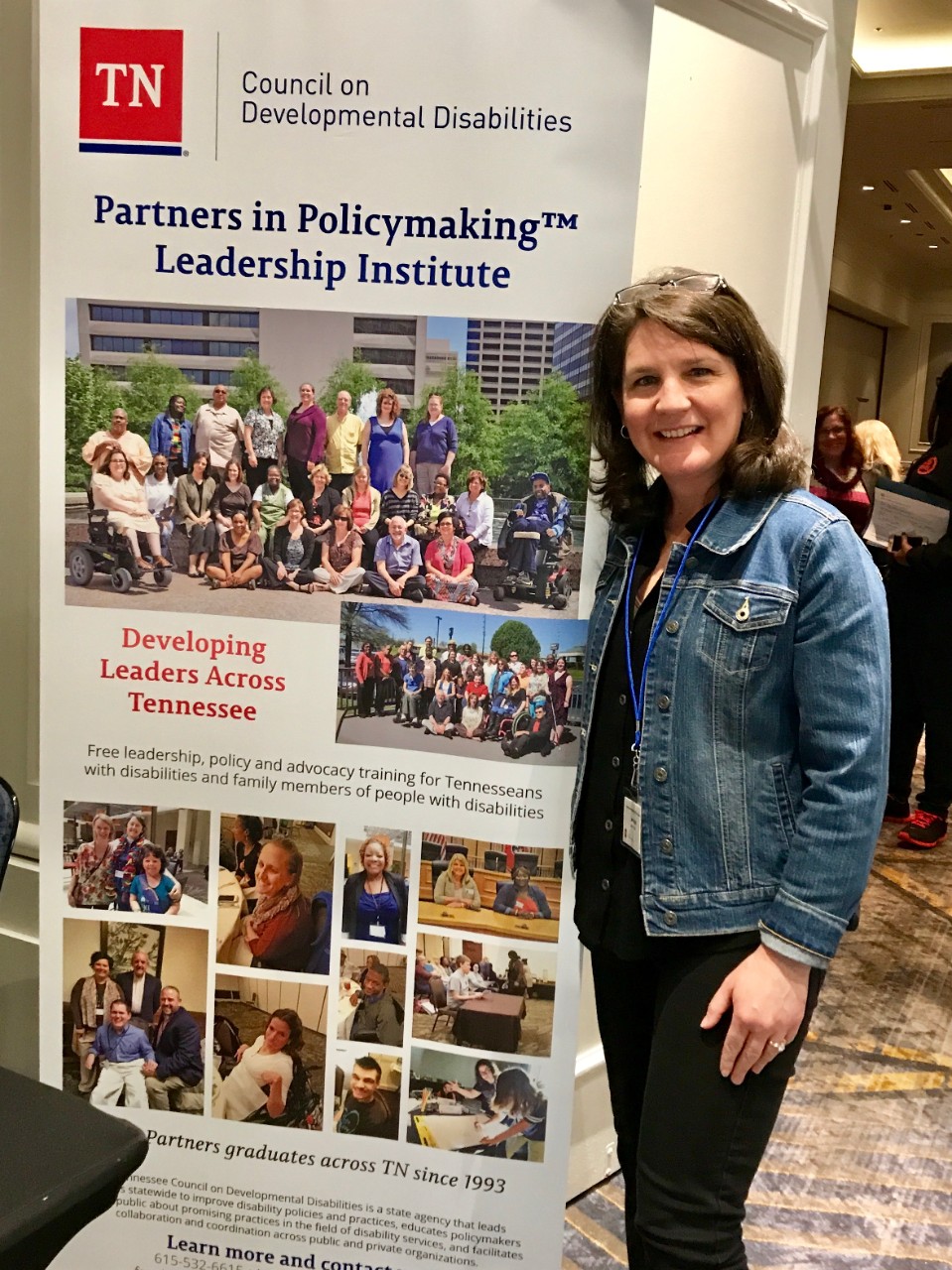 Jen, a woman with wavy, brown hair, poses in front of a Partners banner. She has reading glasses on her head and is wearing a denim jacket and an event badge around her neck. 