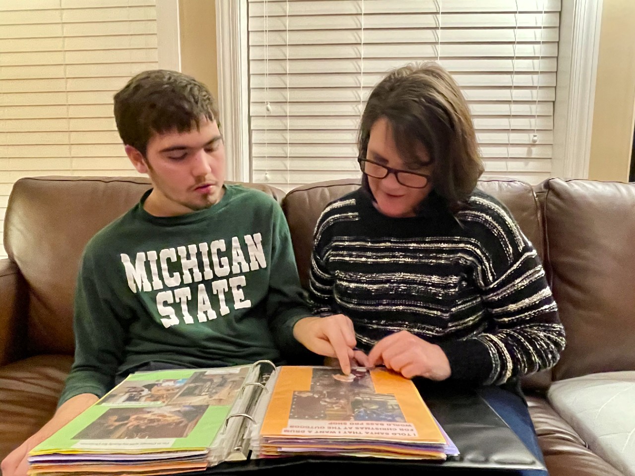 Jen is wearing a striped sweater and is seated on a brown couch next to a young man with brown hair and beard, wearing a green Michigan State shirt. He is pointing to a photo in a binder.  