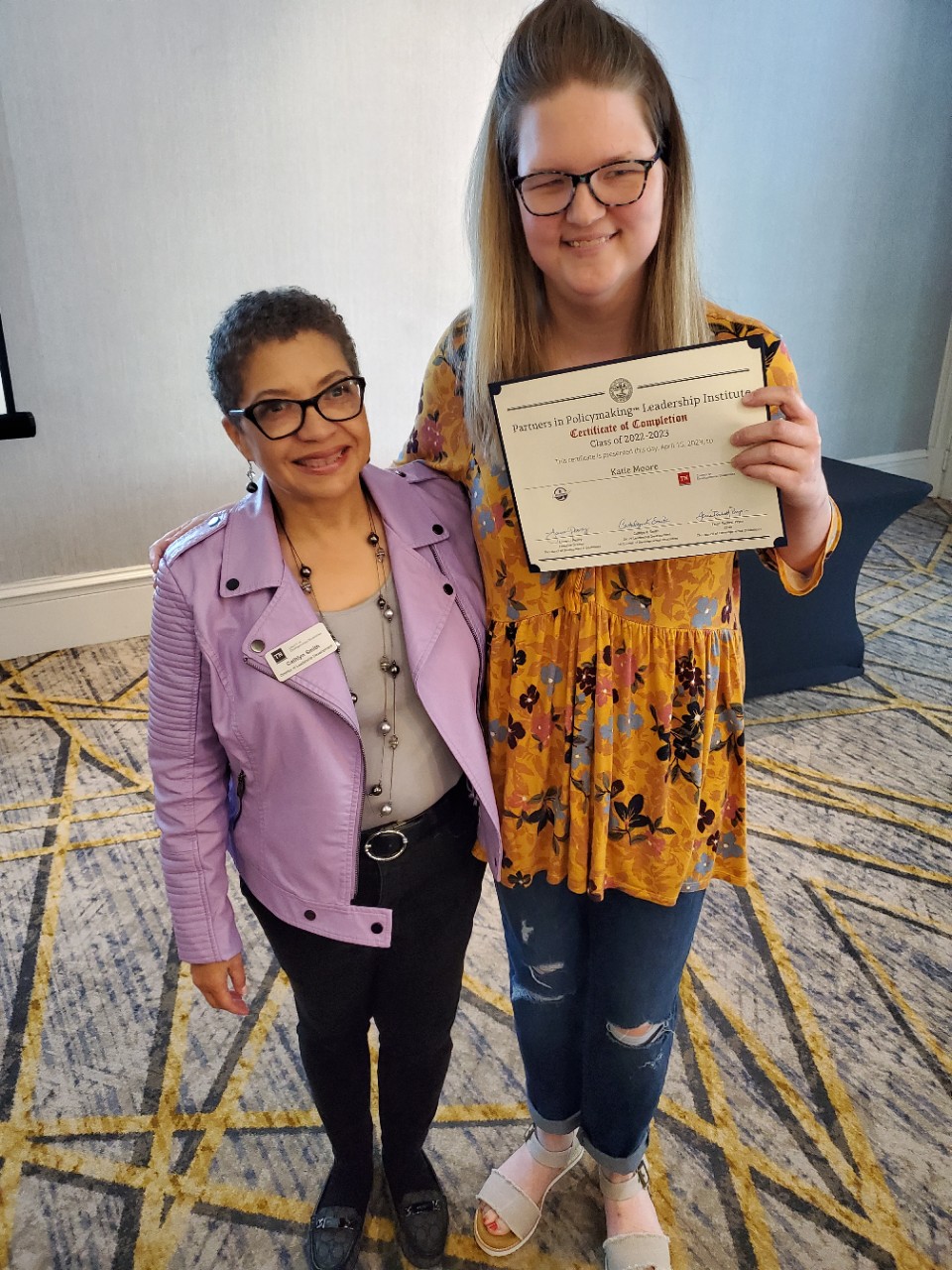 Cathlyn Smith stands with her arm around Katie, a tall, blonde young woman in glasses and a yellow floral top and jeans, smiling and holding a certificate. 