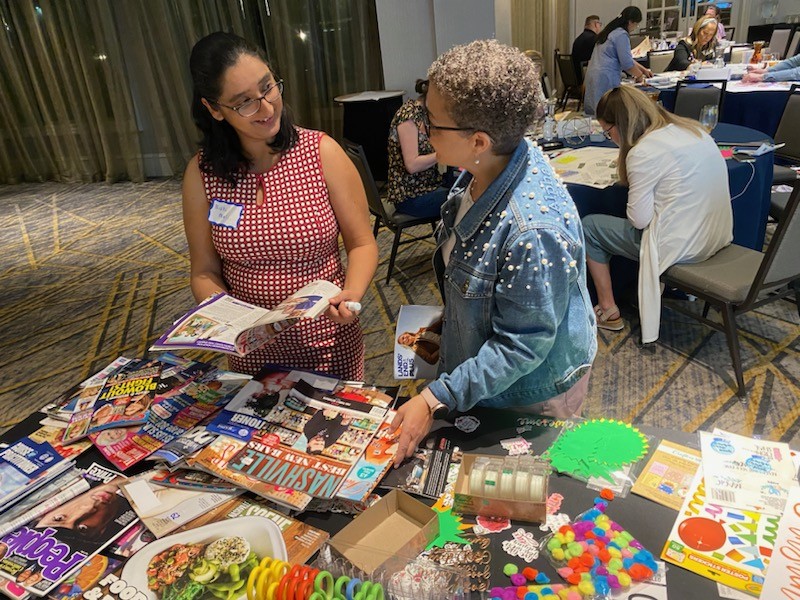 Cathlyn stands at a table covered in craft supplies and old magazines, talking with a woman with dark hair and glasses who is holding an open magazine.  