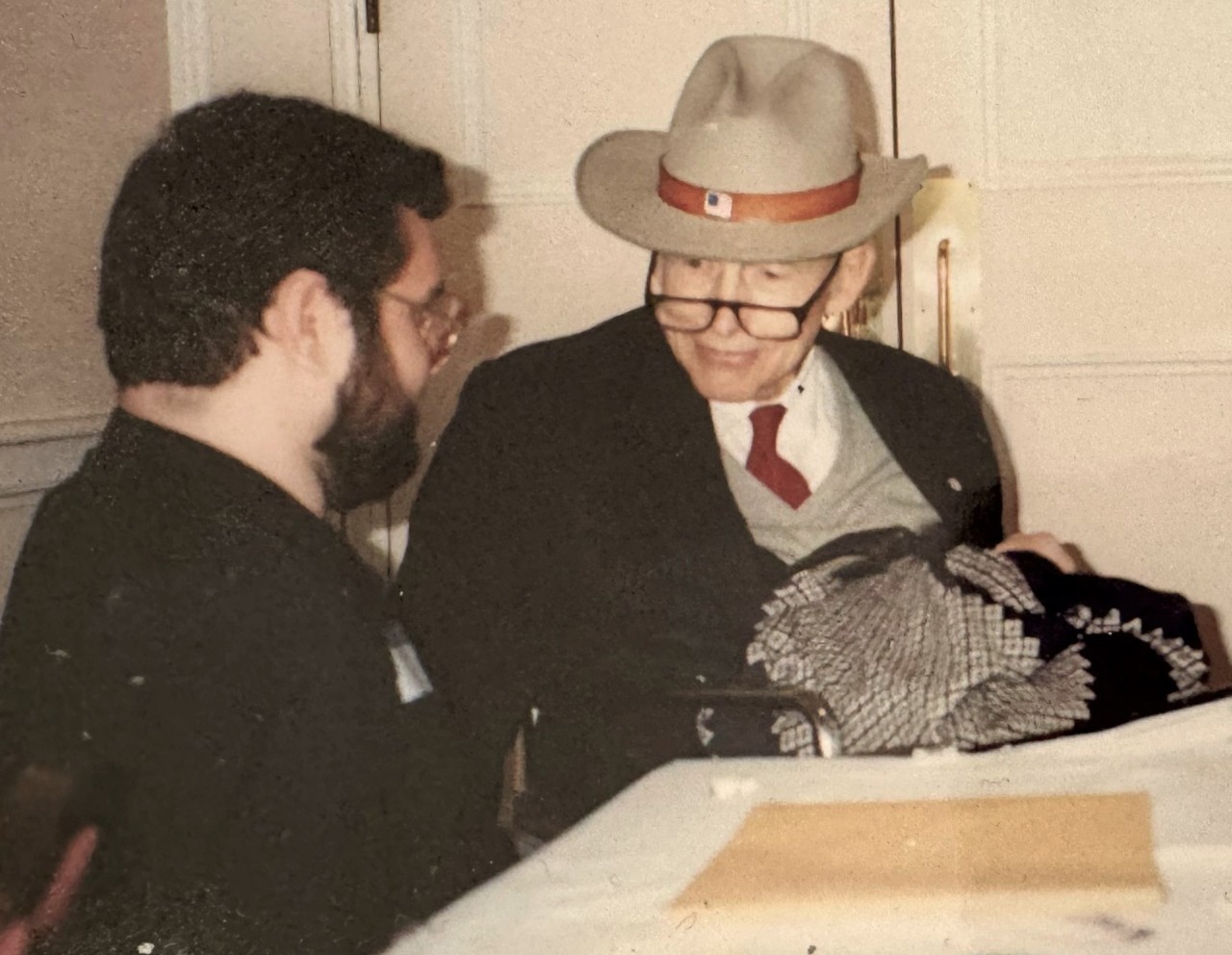 A banner photo at the beginning of the article shows a young, white man with dark hair and beard and glasses seated with an older, white man wearing a tan hat, reading glasses, and a tie, sweater vest, and suit jacket. 