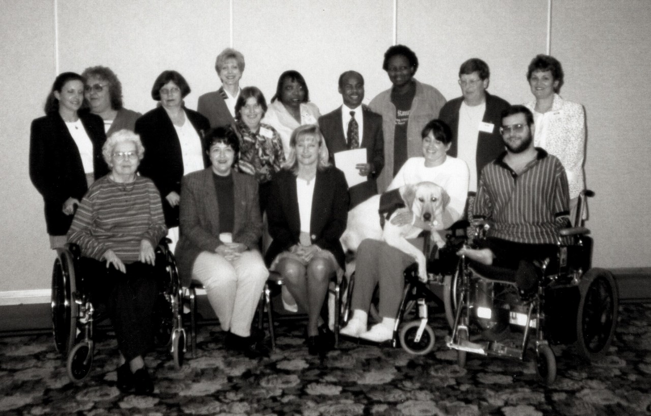 a black-and-white photo of a posed group of 15 people, some in wheelchairs, one with a service dog