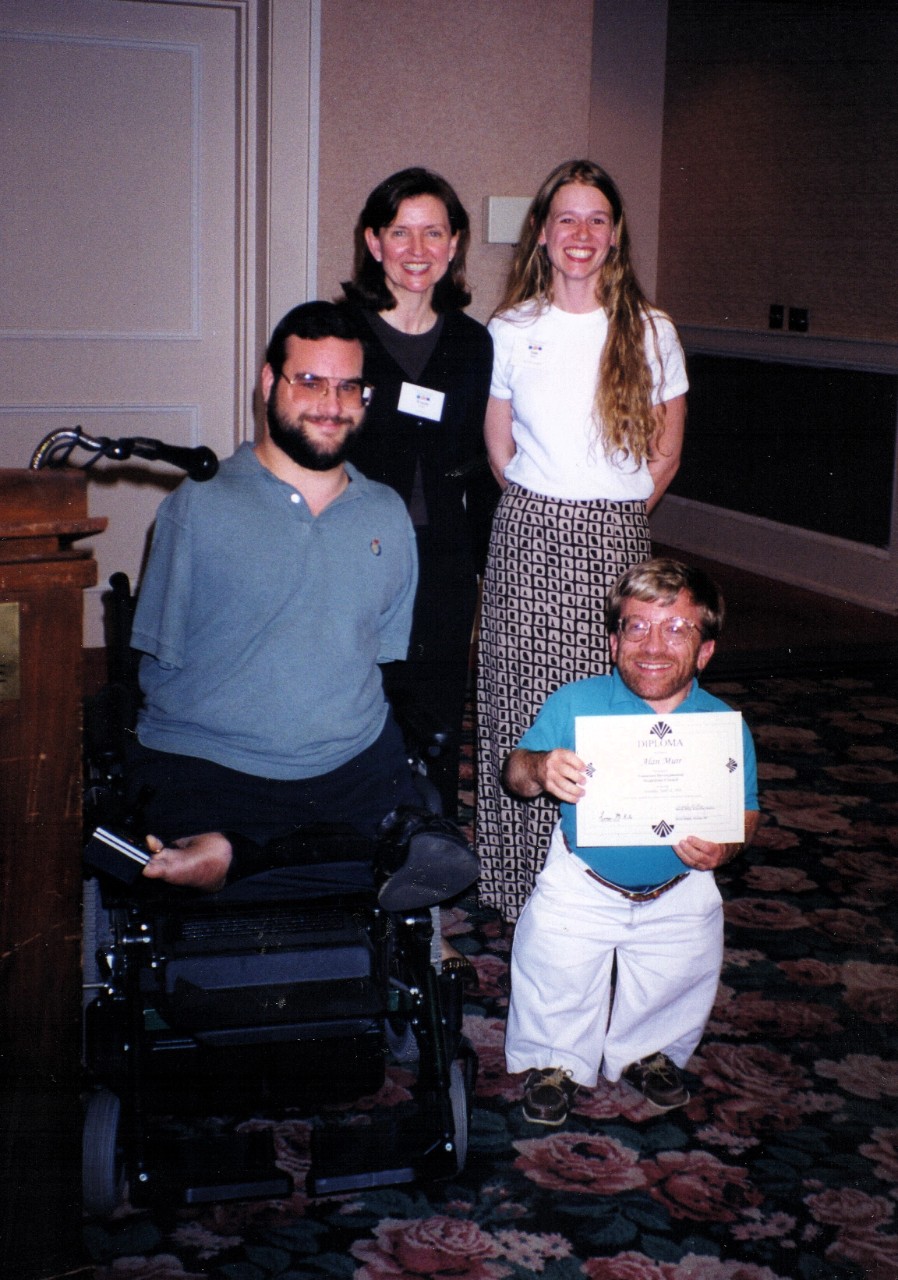Two white women, a white man who is a small person and is holding a certificate, and Kevin Wright, who is using a wheelchair and has limb differences, smile at the camera.