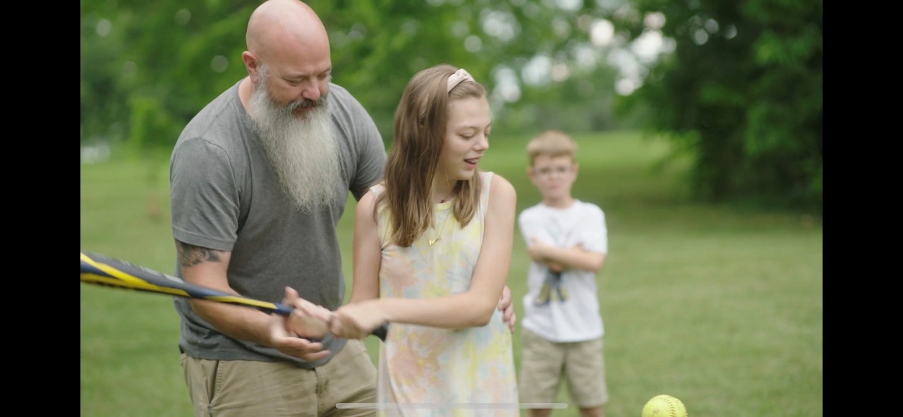 a still shot from the documentary, showing an adult man with a gray t shirt and white beard standing behind a girl of about 10 or 11 helping her swing a baseball bat. the girl is in a white dress and has a visible disability. a young boy looks on from the background. they're all outdoors