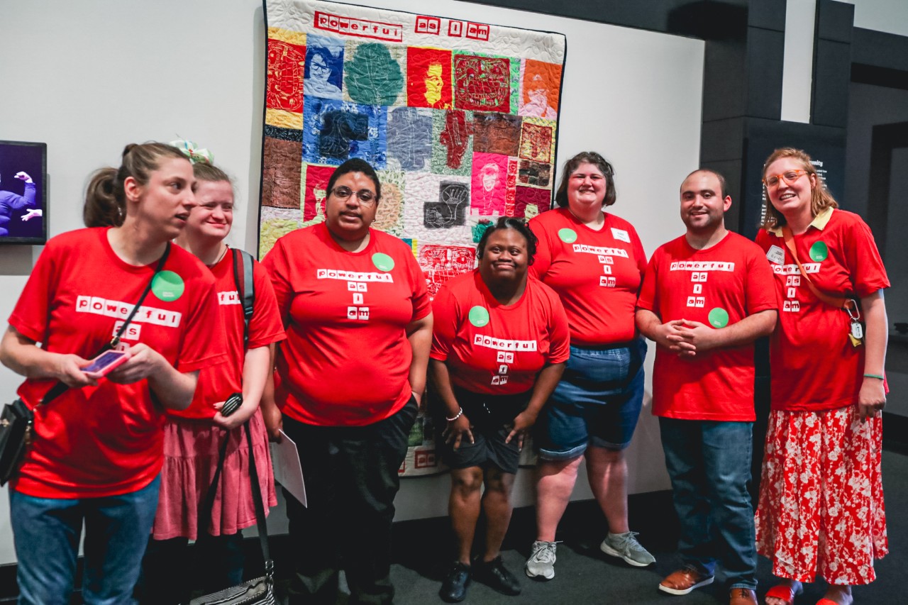 a group of young adults with various disabilities in matching red T-shirts in front of an art gallery wall. Photo credit: Frist Art Museum. 