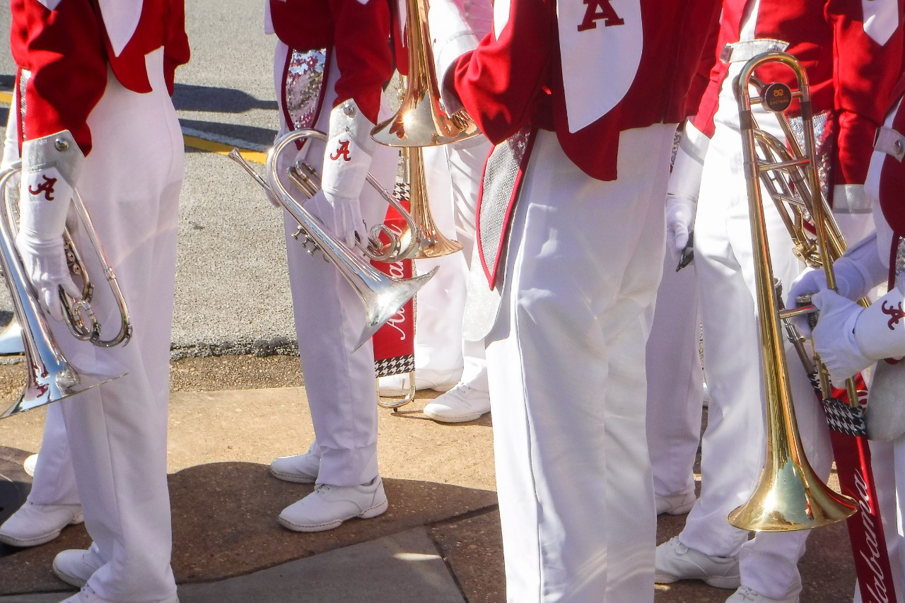 A photo shows brass instruments being held by red-and-white uniformed University of Alabama marching band members. 