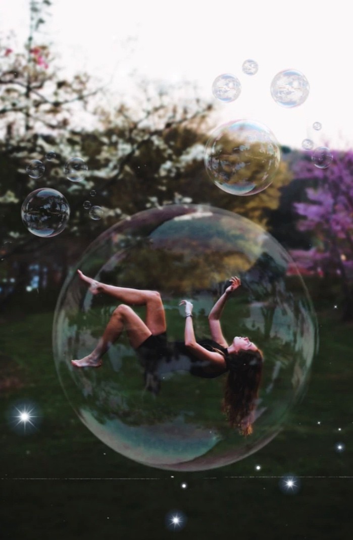 A photo shows large bubbles floating against a background of grass, blooming trees, and a wooded hill. A young woman floats gracefully within the largest bubble, her wavy, dark brown hair hanging down as she is suspended with her legs and arms bent. She is wearing black shorts and a black tank top. 