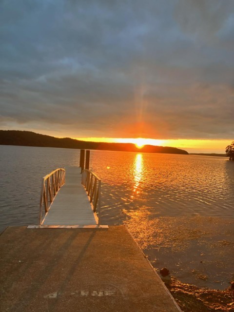  A photo shows a dock and pier extending out into a lake, the water gently rippled. The sun is just slipping behind the hilly shoreline opposite, casting a golden beam of light up into a cloudy sky and down onto the water’s reflection.  The sky beneath the cloud line is brightly golden. 