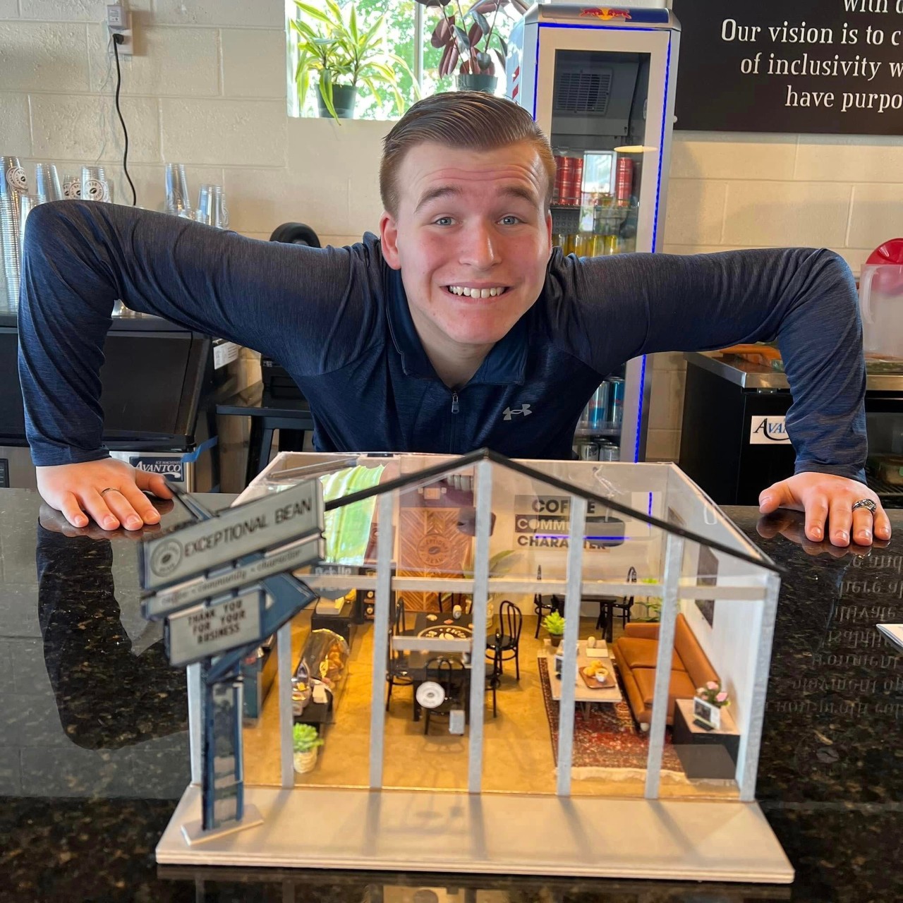 A white, young man with short, sandy-colored hair grins at the camera, eyebrows raised, as he hovers over a miniature building model resting on a countertop. The building is a miniature coffee shop, with a sign out front that reads “EXCEPTIONAL BEAN.” 