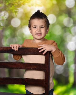 Presley Presley, a young child with a light skin tone and short, spiked brown hair, is smiling at the camera. He is wearing a long-sleeved shirt with tan and brown sections. He is leaning on a wooden chair with a blurred green, leafy background behind him.