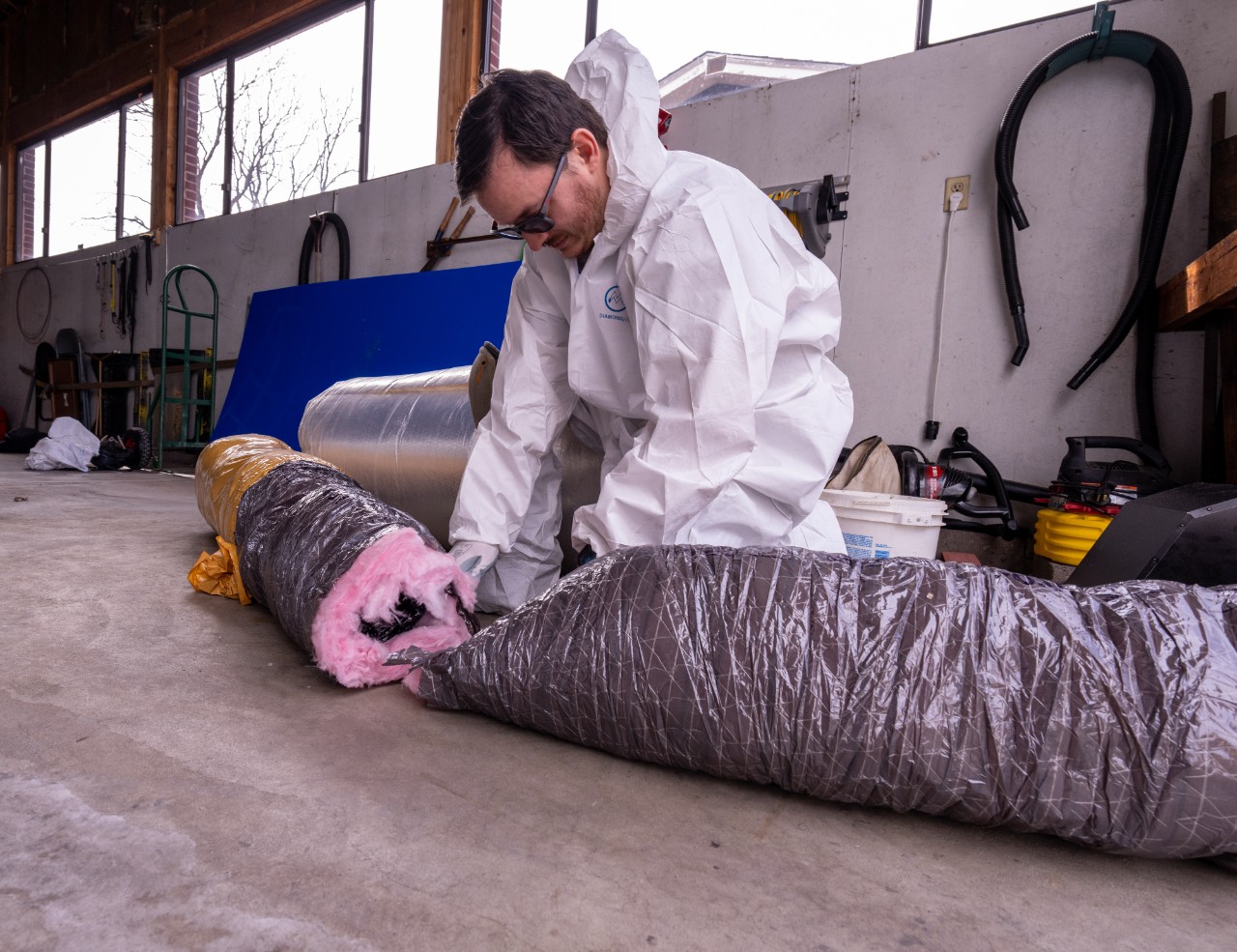 A weatherization crew member from the Energy Resource Center cuts a piece of insulated ducting for a new exhaust fan. | Credit: Werner Slocum / NREL : A weatherization crew member from the Energy Resource Center works in a garage, cutting a piece of insulated ducting wrapped in gray plastic with pink insulation exposed at the end. The worker is wearing a white protective suit, gloves, and safety glasses, focusing intently on the task. Tools and supplies, including hoses, buckets, and a ladder, are visible in the background, along with large windows letting in natural light.