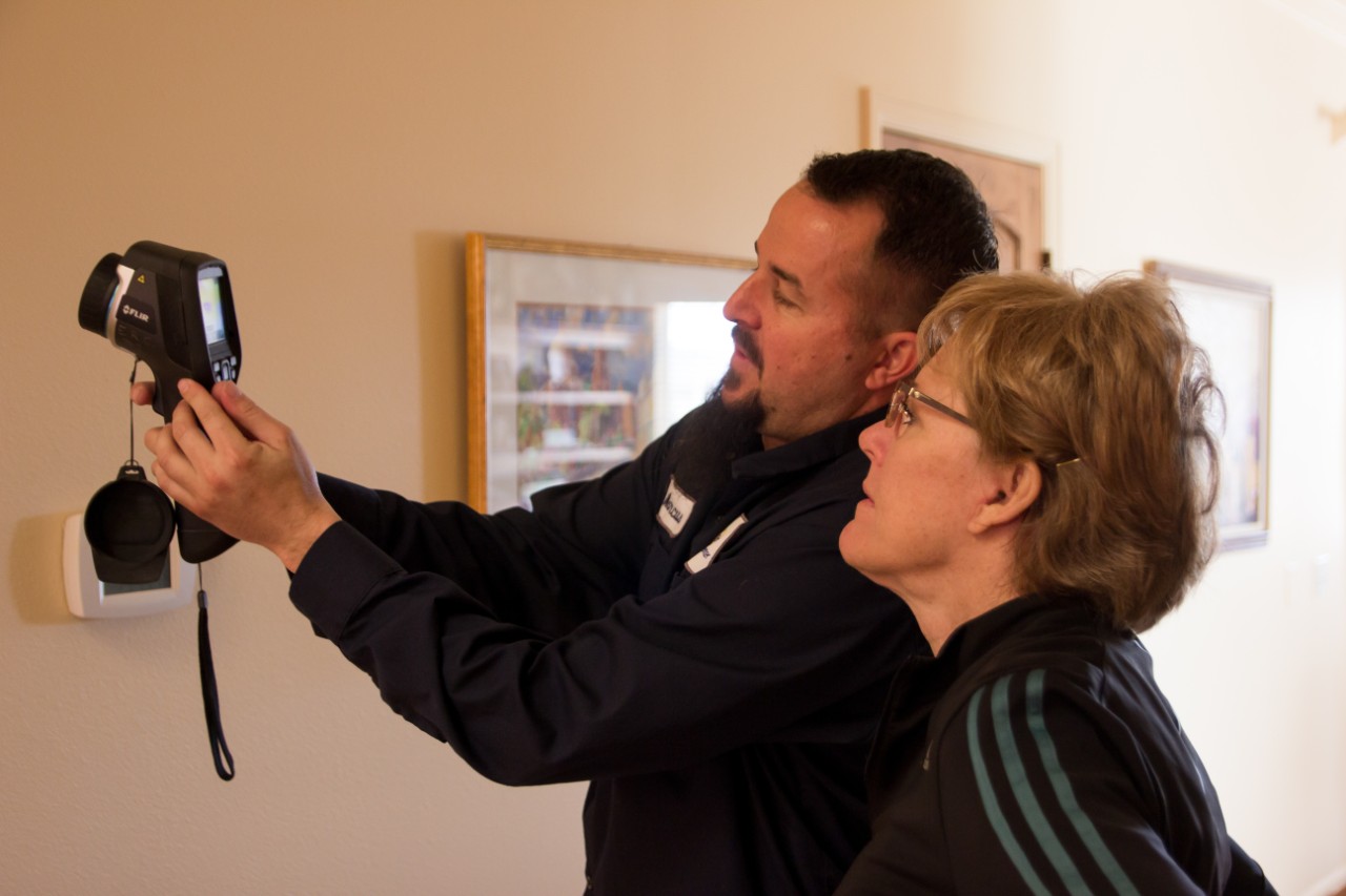 A weatherization technician shows a homeowner an insulation defect identified while using an infrared camera. | Credit: Energy Resource Center A weatherization technician demonstrates an infrared camera to a homeowner, pointing out an insulation defect. The technician, wearing a navy work shirt with a name tag, holds the camera up against a beige wall, focusing on the screen. The homeowner, a light-skinned woman with short hair and glasses, looks attentively at the device. Behind them, framed artwork hangs on the wall, and a thermostat is visible near the camera.