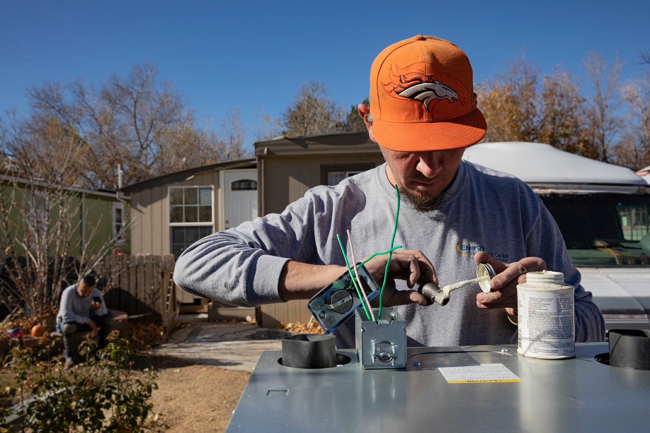 An HVAC technician prepares a gas fitting during a manufactured home furnace replacement. | Credit: Energy Resource Center An HVAC technician works on preparing a gas fitting during the replacement of a furnace in a manufactured home. He wears an orange Denver Broncos cap, a gray long-sleeve shirt with an energy services logo, and focuses intently on his task. Green wires and a metal fitting are visible on the equipment he is working on, alongside a container of sealant. In the background, a manufactured home with a small porch and steps is visible, along with bare trees under a clear blue sky.