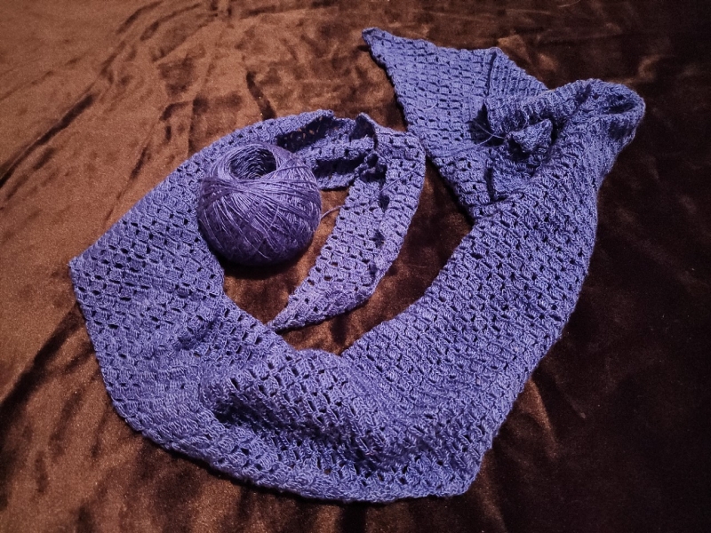 A photograph of a partially completed deep blue crocheted scarf with a lacy, openwork pattern, laid out on a dark, velvety surface. A matching ball of yarn rests near the center, with a strand still connected to the scarf, indicating the piece is still in progress. The scarf has an elegant, lightweight texture with pointed edges, and its intricate stitchwork is clearly visible. 