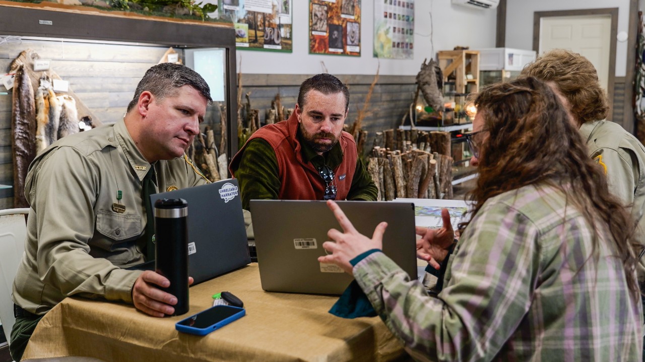 In a nature-center office, Ryan Jolley sits at a table in a rust-red fleece vest and looks at a laptop. A uniformed park ranger sits to his left holding a travel mug, while a person in a plaid shirt gestures toward another laptop on the right. There is a second ranger is partially visible. Fur pelts, wildlife posters, and wooden displays line the background.