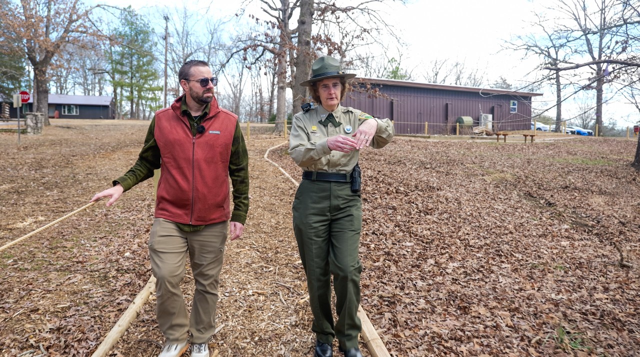 Ryan Jolley walks beside a uniformed park ranger who gestures while speaking, outdoors on a winter day. They follow a wood-chip path bordered by a rope handrail, with leaf-covered ground, bare trees, and a brown building in the background.