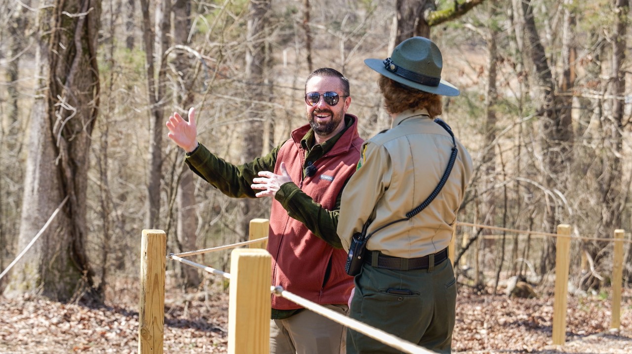 : Ryan Jolley, talking with a uniformed park ranger on a wooded path. He wears mirrored sunglasses and a rust-red fleece vest over an olive shirt, smiles, and gestures broadly as wooden posts with a rope guide the path among leafless trees.