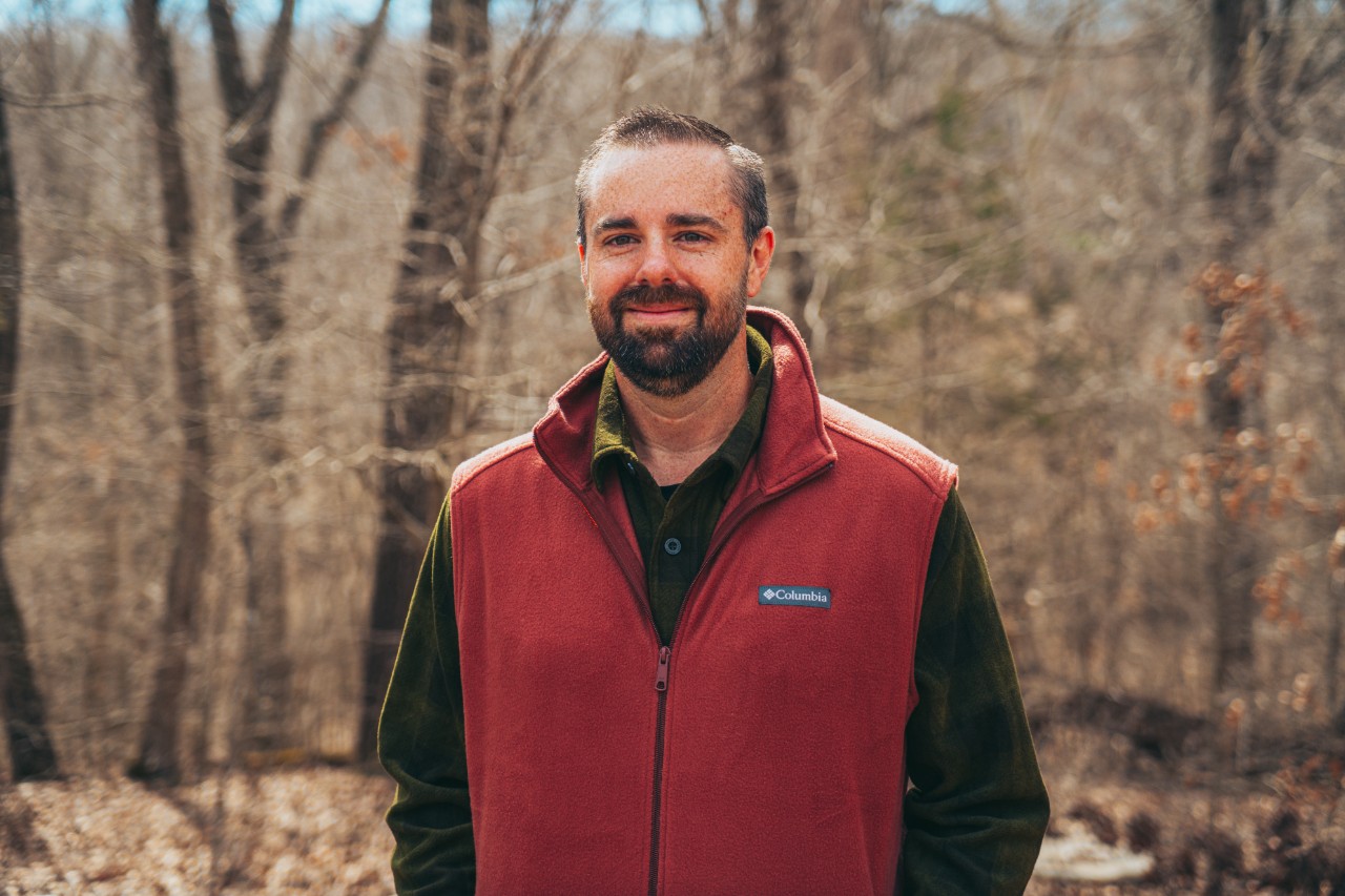 Ryan Jolley stands outdoors in a woodland with leafless trees and soft light. He has light-to-medium skin, short dark hair, and a neatly trimmed beard, and he smiles gently at the camera. He wears an olive-green shirt under a rust-red Columbia fleece vest.