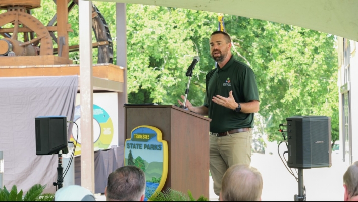 Ryan Jolley speaking at an outdoor podium with a Tennessee State Parks emblem. He has light-to-medium skin, short dark hair, and a beard, and he wears a dark green polo and khaki pants while addressing an audience. A large historic mill wheel and bright green trees form the background, with speakers and equipment visible around the stage.