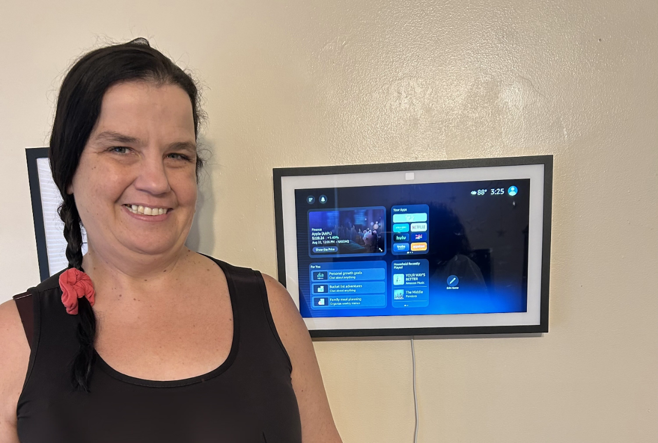 Vicki Kee, a light-skinned woman with dark hair in a braid tied with a red scrunchie, stands smiling beside a mounted smart display on a wall. The screen shows a home interface with various apps and options. Vicki is wearing a black tank top and patterned skirt.