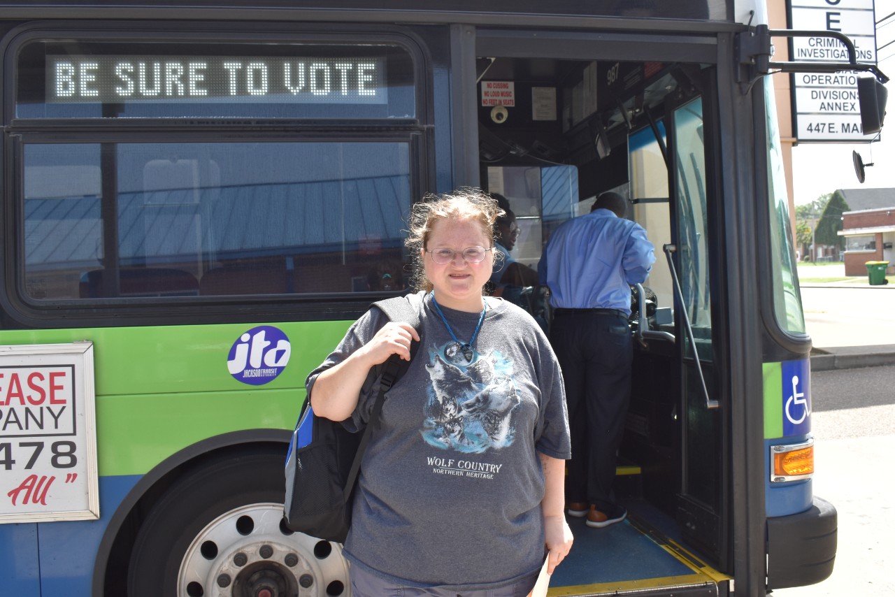 Jennifer Storey, a light-skinned woman with glasses and curly hair pulled back, stands smiling in front of a green and blue public bus. She's wearing a grey "Wolf Country" T-shirt and carrying a backpack over one shoulder. The digital sign on the bus reads “BE SURE TO VOTE,” and the bus driver is visible in the background.
