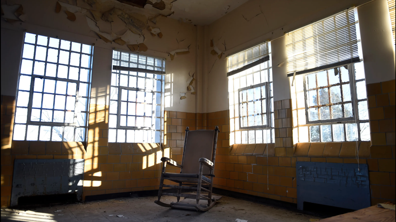 clover bottom Harsh winter light streams through tall, barred windows into a decaying room. Paint peels from the walls above mustard-colored tiles. A single wooden rocking chair sits in the center, casting a long shadow across a dusty floor. The scene evokes abandonment and the institutional past of the Clover Bottom Developmental Center.